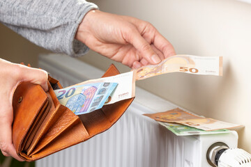 Woman's hand takes euro money banknotes from wallet and places on heating radiator battery with...
