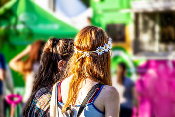 Obraz na płótnie Canvas Two girls - one with red hair and daisy headband - wait in line at food truck at carnival - bright colors of pink and green
