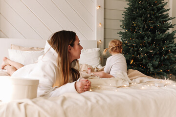 Obraz na płótnie Canvas A young mother and a sad, touchy child are sitting on a bed in a bedroom with a Christmas tree in a cozy house waiting for the Christmas holidays and New Year. Family education. Selective focus