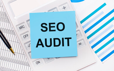 On the desktop is a white calculator with a blue sticker with the text SEO AUDIT, a pen and blue reports. Business concept