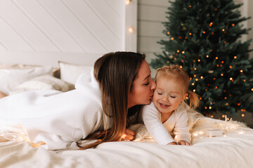 Obraz na płótnie Canvas Cute mom and little daughter baby in pajamas laughing smiling spending time together sitting on the bed in a cozy decorated bedroom with a Christmas tree in the New Year holiday at home. Selective foc