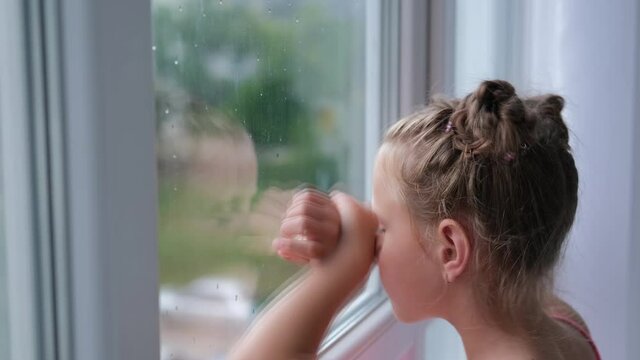 The girl stands at the window and looks at the rain. The child is sad at the window.