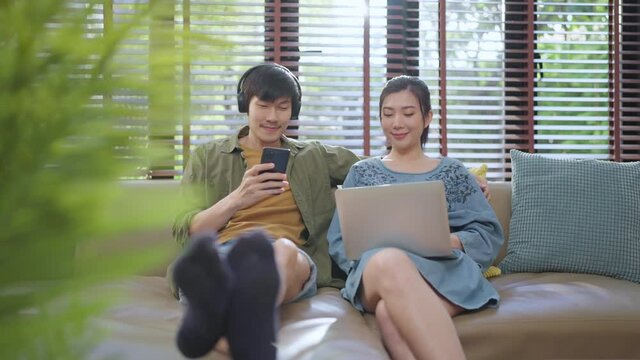 lovely asian marry couple adult stay home lifestyle male playng game smartphone online with his wife smile laugh use laptop device sit relax together on sofa living room at home stay home concept