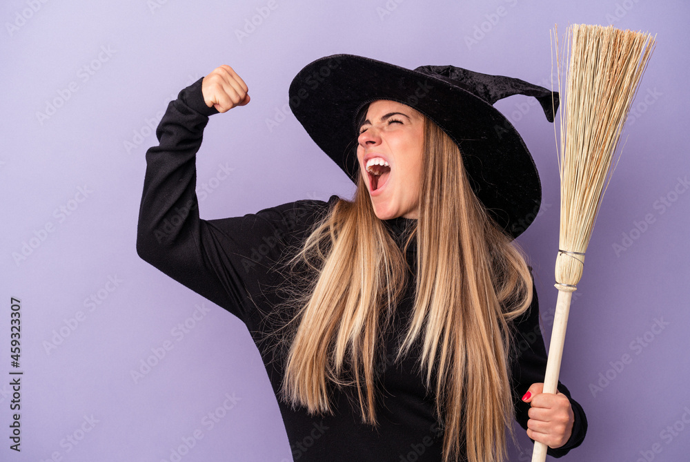 Wall mural young russian woman disguised as a witch holding a broom isolated on purple background raising fist  - Wall murals