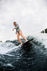Flexible woman in grey swimsuit energetically balancing on wave on wakesurf board.