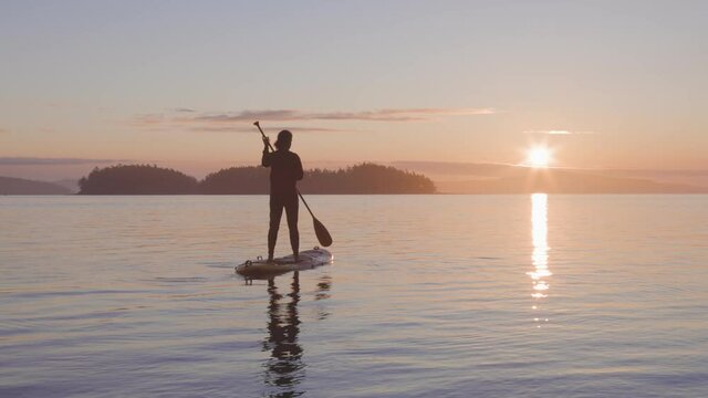 Adventurous Caucasian Adult Woman on a Stand Up Paddle Board is paddling on the West Coast of Pacific Ocean. Sunny Sunrise. Victoria, Vancouver Island, BC, Canada. Slow Motion