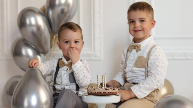 Two boys celebrating birthday, children applauding. Birthday cake with candles and balloons. Happy kids, celebration, white minimalist interior.