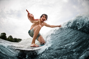 cheerful man show hand gesture riding on surf style wakeboard and touching the wave with one hand