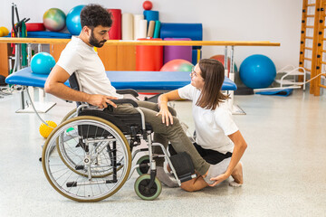 Fototapeta na wymiar Female physiotherapist and male patient seated in a wheelchair during rehabilitation treatment - joint mobility, and strengthening of leg muscles