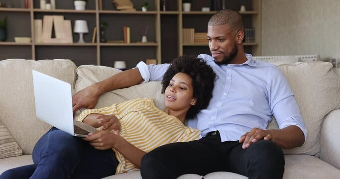 African couple relaxing on couch in living room with laptop, carefree spouses discuss purchase on internet, make order, buy remotely using e-commerce retail virtual services. Modern tech, fun concept