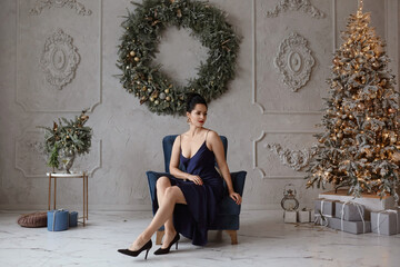Adorable woman in a stylish dark-blue dress in front of Christmas tree. Lovable female model in trendy dress posing in the interior decorated for New year holidays - obrazy, fototapety, plakaty