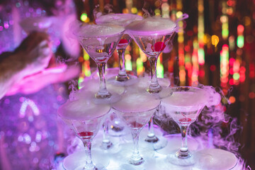Pyramid of glasses with alcoholic beverages, filling and pouring beautiful pyramid line of different colored alcohol cocktails with champagne and dry ice on a party, catering banquet table