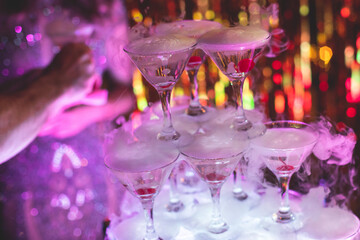 Pyramid of glasses with alcoholic beverages, filling and pouring beautiful pyramid line of different colored alcohol cocktails with champagne and dry ice on a party, catering banquet table