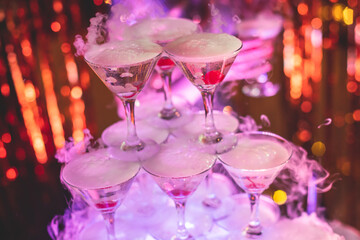 Pyramid of glasses with alcoholic beverages, filling and pouring beautiful pyramid line of different colored alcohol cocktails with champagne and dry ice on a party, catering banquet table