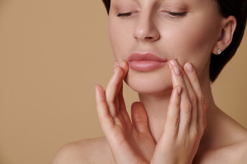 Close-up beauty portrait of pretty woman with clean fresh skin touching her face , doing a rejuvenating massage with fingers. Beauty treatment, skincare and spa concept on beige background, copy space