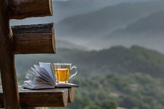 Open book and cup herbal tea on background of foggy mountain forest on wooden rustic bench. Wellness holiday, vacation, healthy rest time in nature Rhodope mountains, Bulgaria
