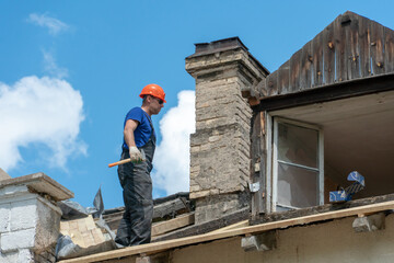 Repair of a wooden roof outdoors against the background of blue sky and clouds. A fashionable carpenter in sunglasses and special clothes with a hammer in his hand installs beams and boards.