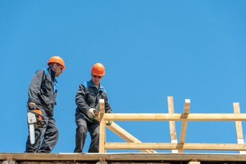 Repair of a wooden roof outdoors on a summer day against the background of blue sky and clouds. A carpenter in special clothes and with a tool installs beams and wood boards.