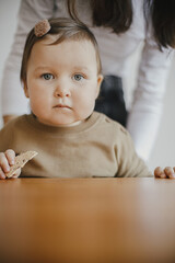 Adorable baby girl holding healthy cookie and sitting at wooden table. Cute toddler girl in modern brown outfit and with hair pin having snack in room