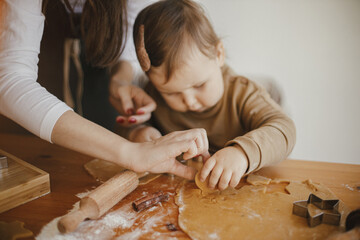 Obraz na płótnie Canvas Adorable little daughter and mother making together gingerbread cookies on messy wooden table in modern room. Cute toddler girl helps making christmas cookies. Mommy daughter lovely moments