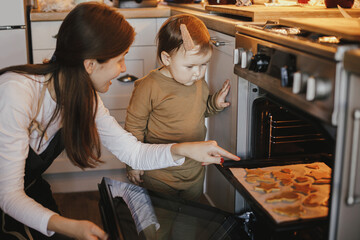 Cute little daughter and mother baking christmas gingerbread cookies in modern scandinavian kitchen. Cute toddler girl and mom putting tray with cookies in oven. Family holiday preparations