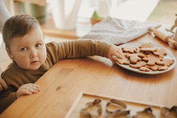 Cute little girl grabbing freshly baked gingerbread cookie on wooden table in modern room. Adorable toddler girl making christmas cookies with mother. Authentic lovely moment, holiday preparation