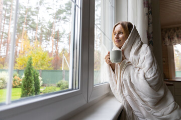 Pensive young woman in sweater drinking hot purifying herbal tea at home. Thoughtful girl drinking coffee, thinking, relaxing, looking out the window, blurry autumn landscape outside. Fall season. 