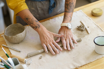 Close up of potter woman rolling piece of clay with her hands on wooden table in pottery, has tattoos on both arms, top view. Female ceramist in workshop during work. Small business, hobby concept.