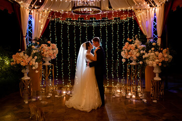 newlyweds at evening wedding ceremony in park among light bulbs and garlands.
