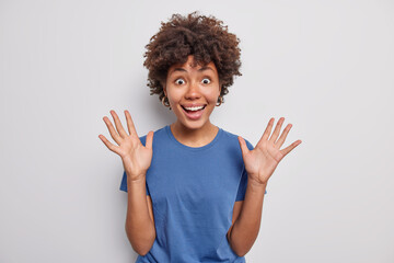 Positive dark skinned ethnic young woman raises palms smiles broadly reacts joyfully at something funny expresses good emotions dressed casually poses against white background. Emotions concept