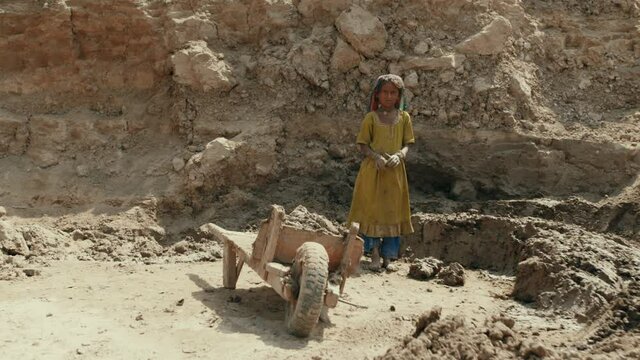 A little girl is in labor slavery in Pakistan. A dirty and barefoot girl in national clothes stands next to a cart against the background of a clay mountain and stones. Kids kidnapping. Full HD