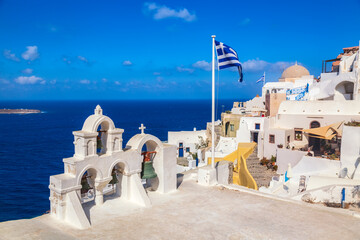 Fototapeta premium Greek orthodox church with bells and greek flag against famous white houses on Santorini island, Aegean sea, Greece.