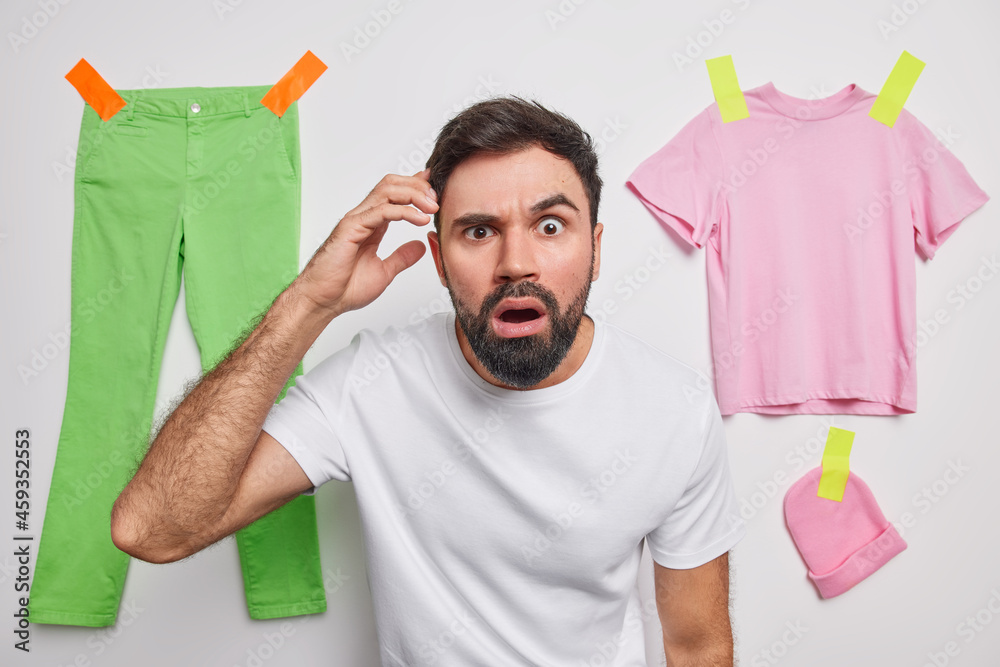 Wall mural Shocked bearded adult man scratches head stares surprised at camera cannot decide what to do dressed in casual t shirt poses against white background with plastered clothes. Oh no whats this