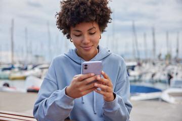 Lovely African American woman with curly hair dressed in sweatshirt holds mobile phone has long...
