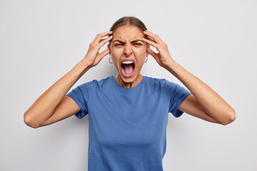 Outraged young woman keeps hands on head yells angrily keeps mouth wide opened loses control has mental breakdown screams furious wears blue t shirt isolated over white background relieves from stress