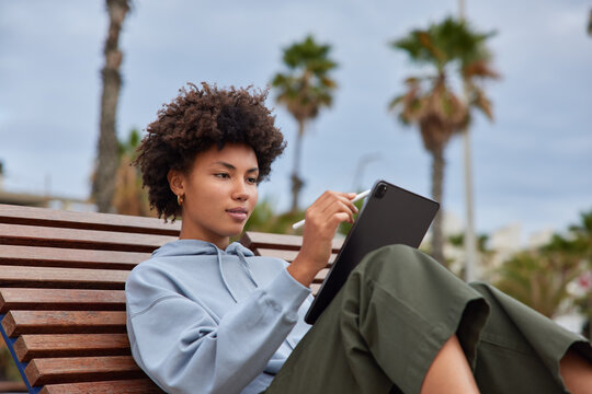 Creative ethnic female graphic designer sketches her project on tablet with stylus poses on wooden bench outdoor works outside. Woman photo editor uses interactive pen display. Skilled retoucher