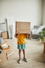 Fototapeta na wymiar Vertical full length portrait of teenage boy with box on head playing with things while family moving to new house