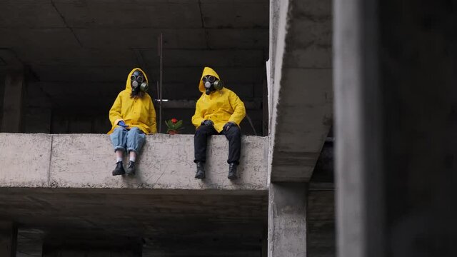 man and woman are sitting on edge of abandoned building in gas masks and yellow protective suits, in middle they have red chrysanthemum flower as symbol of life. Suddenly look in camera