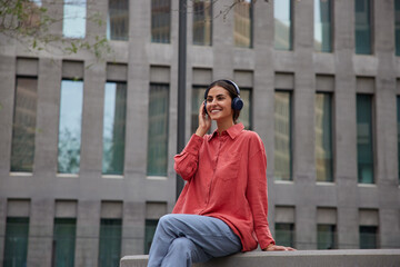 Cheerful beautiful woman sits relaxed against urban setting listens music in wireless headphones dressed in casual shirt and jeans enjoys music playlist popular audio tracks during leisure time.