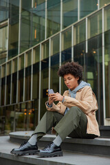 Afro American woman in stylish clothes uses modern mobile phone for chatting online updates application poses outdoor satisfied good internet looks with serious expression poses in urban city