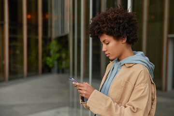 Sideways shot of African American girl writes text message for sending enjoys web browsing for chatting in social networks via cellular application dressed in trendy clothes poses outdoors alone