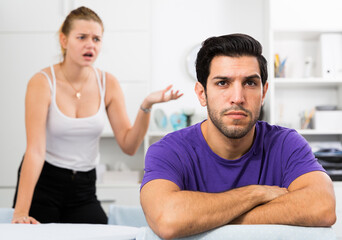 Fototapeta premium Frustrated guy sitting at home table on background with dissatisfied girlfriend
