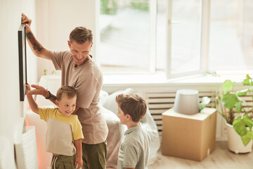 Side view portrait of happy father with two sons hanging pictures on wall while moving in to new...