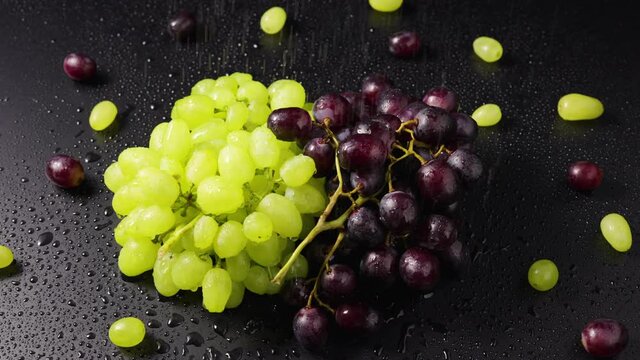 Bunches of green and blue grapes lie on the wet table surface under rain. Top view close up of ripe grape berries rotating in slow motion. Wet berries of juicy grapes. Autumn harvest of sweet fruits.