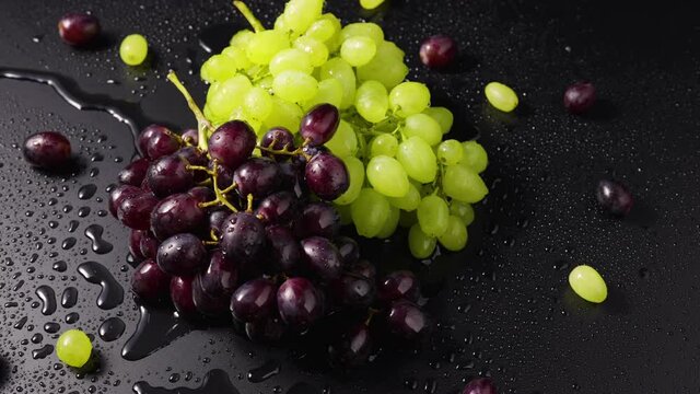 Bunches of green and blue grapes with drops of moisture lie on the wet table surface. Top view close up of ripe grape berries rotating in slow motion. Grapevine in drops of moisture. Sweet fruits.