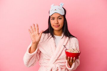 Young latin woman wearing pajama holding a bowl of cereals isolated on pink background  smiling cheerful showing number five with fingers.