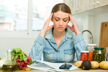 Serious young woman working with papers and using phone at home kitchen