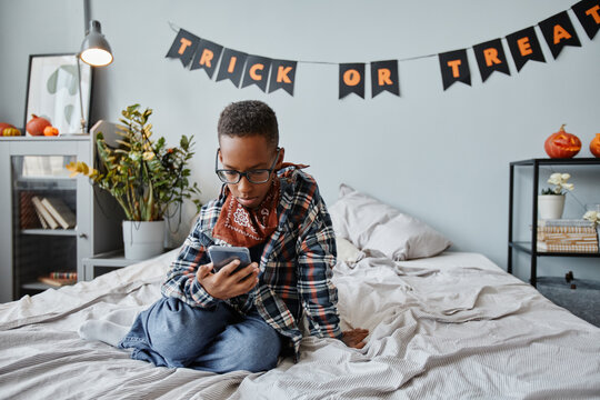 Full length portrait of teenage boy using smartphone on bed in room with Halloween decorations, copy space