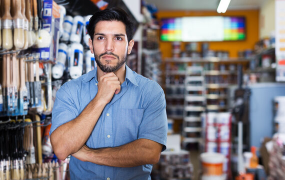 Thoughtful serious young man standing amongst racks with tools in paint store