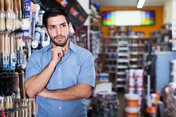 Thoughtful serious young man standing amongst racks with tools in hardware store